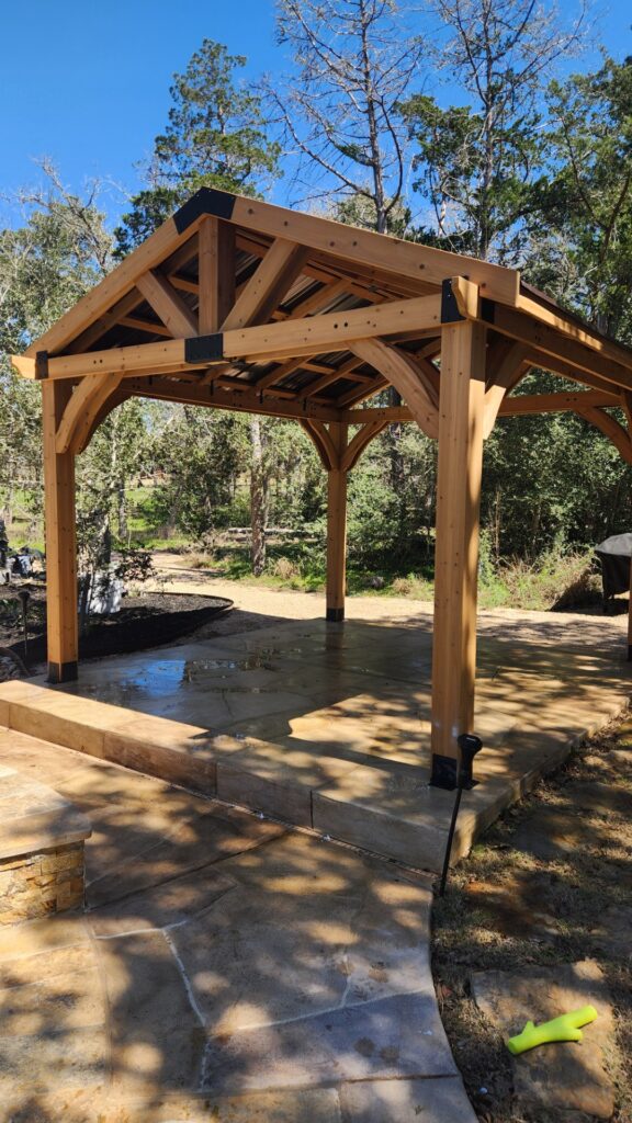 A wooden gazebo structure in a natural outdoor setting with stone pathways and surrounding trees.